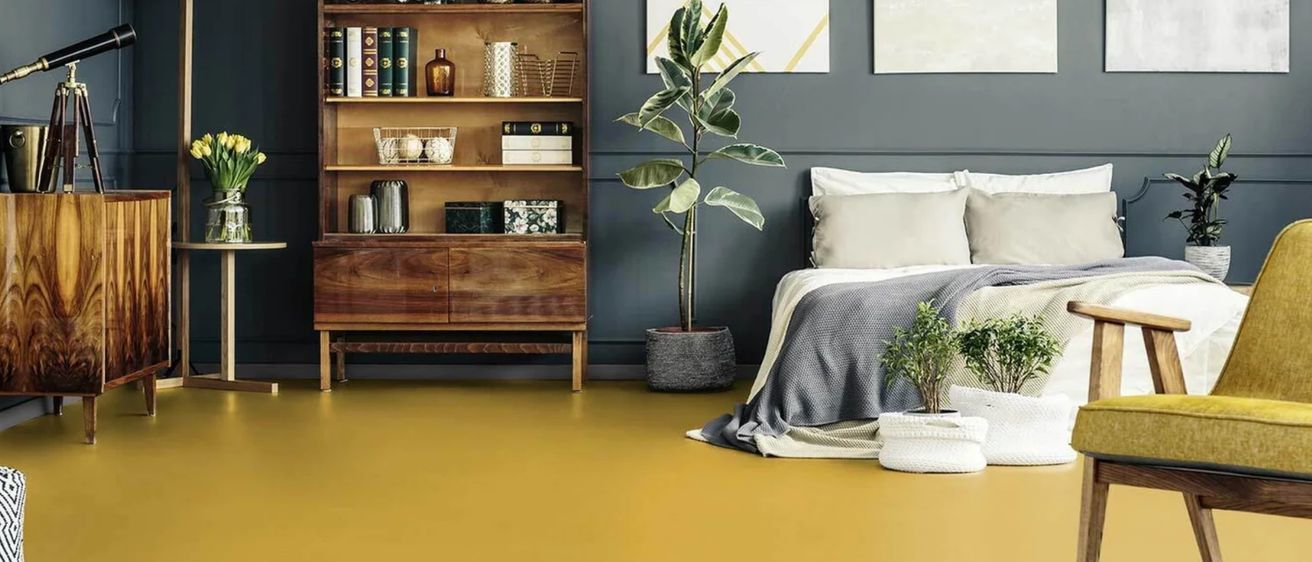 Ein Schlafzimmer mit Bett, Kissen und Decken. Ein Holzschrank mit Büchern und Pflanzen darauf. Ein Topf mit Pflanzen steht auf dem Boden. Drei gerahmte Kunstwerke hängen an der Wand.