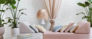 Ein Wohnzimmer mit weißer Ziegelwand und einem rosa Sofa mit mehreren Kissen. Ein Topf mit Pflanze und ein Glastisch sind in der Nähe. Eine Lampe und ein Vorhang ergänzen die Einrichtung.