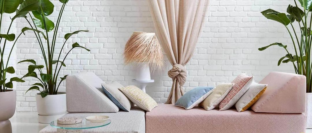 Ein Wohnzimmer mit weißer Ziegelwand und einem rosa Sofa mit mehreren Kissen. Ein Topf mit Pflanze und ein Glastisch sind in der Nähe. Eine Lampe und ein Vorhang ergänzen die Einrichtung.