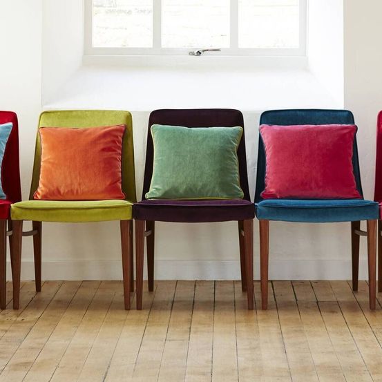 Vier Holzstühle mit Samtkissen in Orange, Grün und Rot sind in einer Reihe vor einem Fenster aufgestellt.
