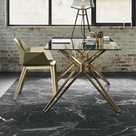 Ein modernes Büro-Setup mit einem Glastisch mit Messingbeinen und einem grünen Stuhl vor einer Ziegelwand mit Fenster. Der Tisch enthält eine Lampe, Bücher und Büromaterial.