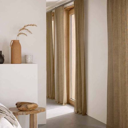 Ein minimalistisches Schlafzimmer mit einem Holztisch, einem Steinobjekt, einer Vase mit getrockneten Gräsern und beigen Vorhängen. Der Raum hat weiße Wände und einen Holzfensterrahmen.