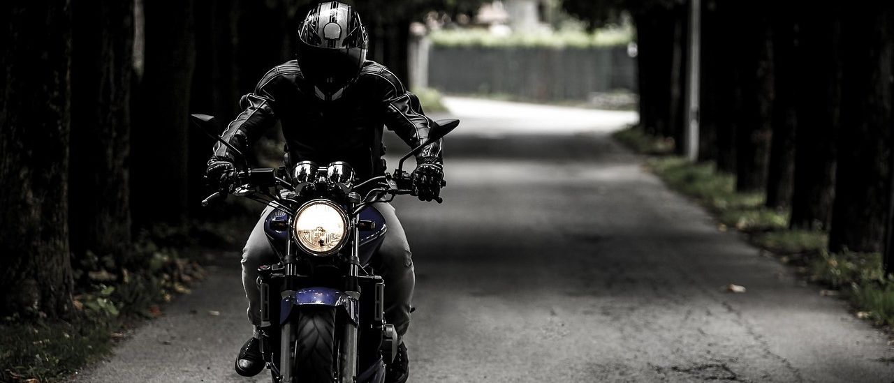 Ein Motorradfahrer fährt mit einem blauen Motorrad auf einem dunklen, gewundenen Weg. Er trägt einen Helm, Handschuhe und eine Lederjacke. Das Scheinwerferlicht des Motorrads erhellt den Weg vor ihm.