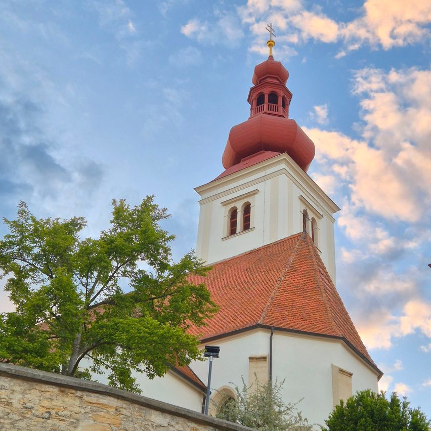 Eine Kirche mit einem roten Turm und einer Spitze steht vor einem blauen Himmel mit weißen Wolken. Ein Baum steht vor der Kirche, und in der Nähe befindet sich eine Straßenlaterne.