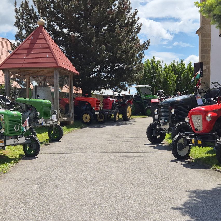 Eine Gruppe von Oldtimer-Traktoren auf einem Einfahrtsweg geparkt, mit einem roten Pavillon und einem Gebäude im Hintergrund, unter einem teilweise bewölkten Himmel.