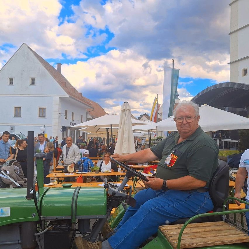 Ein älterer Mann fährt einen grünen Traktor auf einem Stadtplatz, mit einem Außencafé im Hintergrund unter einem bewölkten Himmel.