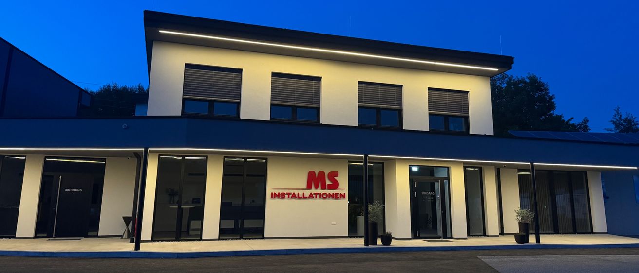 Ein modernes Bürogebäude mit einem roten Schild, auf dem 'MS Installationen' steht. Das Gebäude hat eine weiße Fassade, Glasfenster und eine Glastür. Es ist nachts beleuchtet.