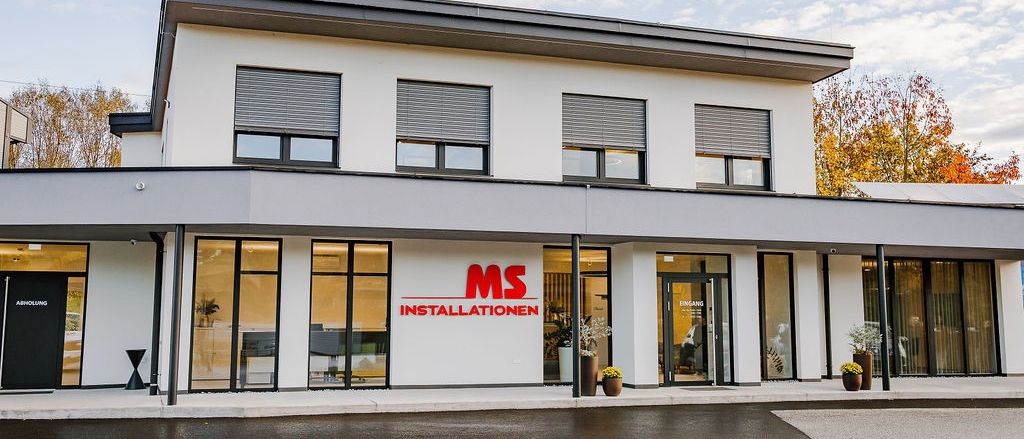 Ein modernes weißes Gebäude mit dem Logo MS Installationen. Es hat Glasfenster, Glastüren und Topfpflanzen an den Seiten.