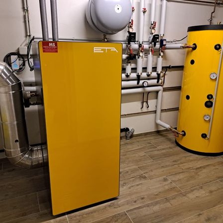 Ein gelber ETA-Wasserspeicher ist in einem Raum mit Holzboden installiert. Ihm stehen ein Wassertank und mehrere an der Wand montierte Rohre und Messgeräte zur Seite.