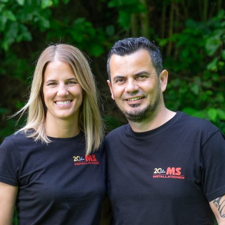 Ein Mann und eine Frau stehen lächelnd eng beieinander vor einem grünen Hintergrund. Sie tragen passende schwarze T-Shirts mit dem Text '20 Jahre MS Installationen'.