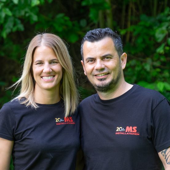 Ein Mann und eine Frau stehen lächelnd eng beieinander vor einem grünen Hintergrund. Sie tragen passende schwarze T-Shirts mit dem Text '20 Jahre MS Installationen'.