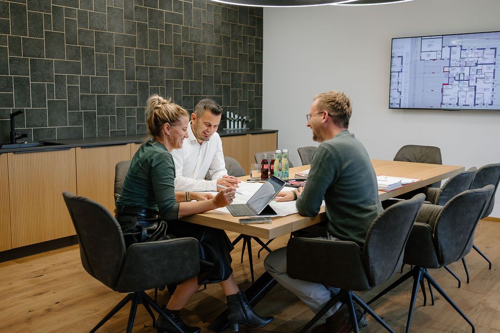 Drei Menschen sitzen lächelnd um einen Schreibtisch. Ein Laptop und Papiere sind auf dem Tisch. Eine Tafel ist an der Wand.