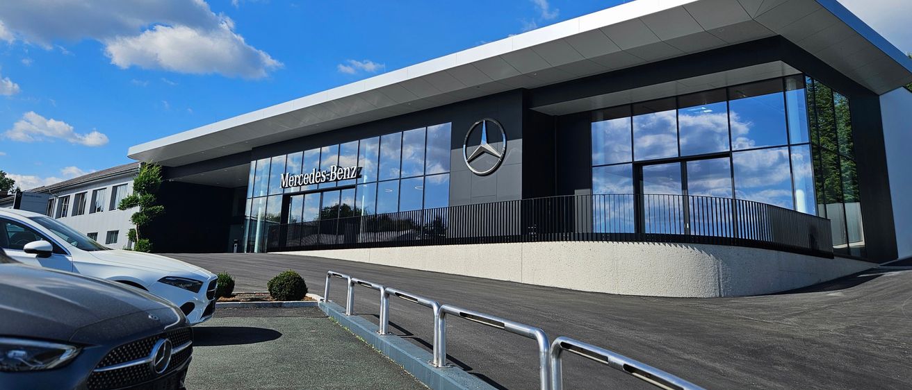 Das moderne Außendesign des Mercedes-Benz-Händlers zeichnet sich durch ein schlankes Design mit Glaswänden, einem großen Mercedes-Benz-Logo und mehreren vor dem Gebäude geparkten Autos aus.