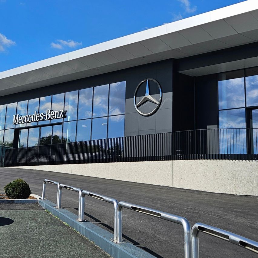 Das moderne Außendesign des Mercedes-Benz-Händlers zeichnet sich durch ein schlankes Design mit Glaswänden, einem großen Mercedes-Benz-Logo und mehreren vor dem Gebäude geparkten Autos aus.