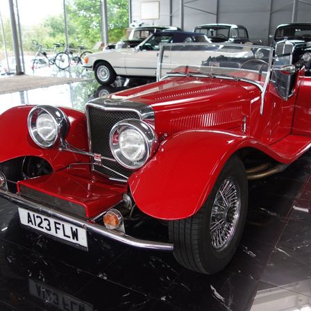 Ein roter klassischer Cabriolet ist in einer Ausstellung geparkt. Das Auto hat das Kennzeichen A123 FLW. Dahinter sind weitere Autos geparkt.
