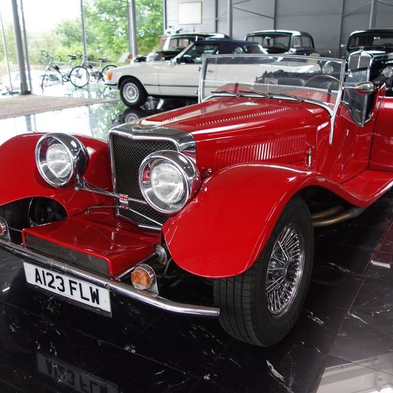 Ein roter klassischer Cabriolet ist in einer Ausstellung geparkt. Das Auto hat das Kennzeichen A123 FLW. Dahinter sind weitere Autos geparkt.