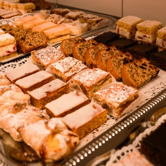 Ein Tablett mit verschiedenen Gebäcksorten, darunter Kuchen, Törtchen und Brote, alles mit Puderzucker bestreut und auf Spitzendeckchen angeordnet.