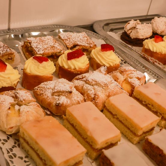 Ein Tablett gefüllt mit verschiedenen Gebäckstücken. Es gibt Sahnetorten, einige mit Erdbeeren darauf, und andere mit Puderzucker bestreut. Das Sortiment umfasst auch geschichtete Desserts mit einer rosa Glasur.