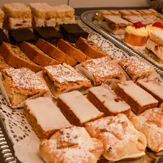 Ein Tablett mit verschiedenen Desserts ist ausgestellt, darunter Brownies, Kuchen und Brotscheiben mit Puderzucker darauf. Jedes Dessert ist ordentlich auf einem weißen Spitzendeckchen angeordnet.