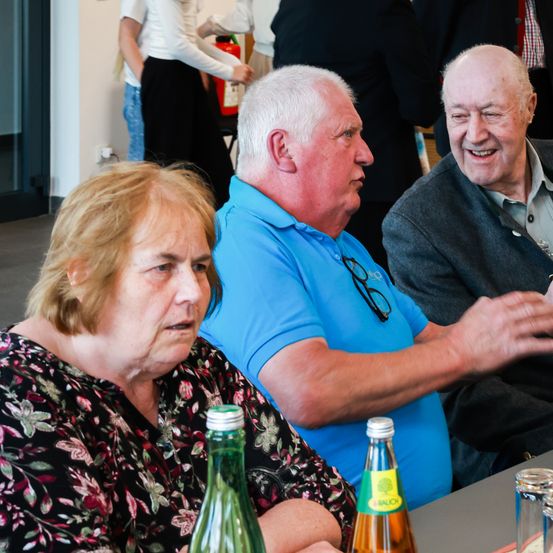 Eine ältere Frau und zwei ältere Männer sitzen an einem Tisch mit Getränken, während andere im Hintergrund herumlaufen.