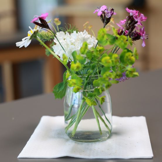 Ein Glas mit Wasser enthält einen Strauß verschiedener bunter Blumen. Die Blumen stehen auf einem weißen Serviettentuch über einem grauen Tisch. Der Hintergrund ist leicht verschwommen.