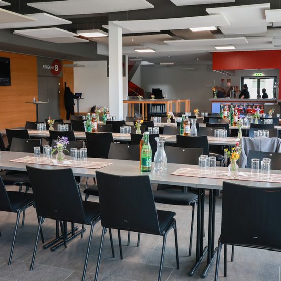 Ein Konferenzraum mit Reihen schwarzer Stühle und Tische. Jeder Tisch hat eine Flasche, eine Vase mit Blumen und Gläser. Eine Person steht auf dem Podium auf der linken Seite.