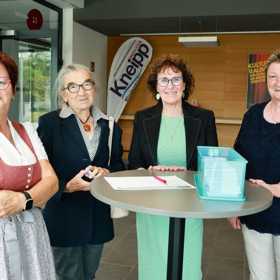 Vier Frauen in Anzügen und Brillen stehen um einen runden Tisch herum, möglicherweise ein Dokument unterzeichnend. Eine Frau hält einen Stift. Ein Aufbewahrungsbehälter und Papiere liegen auf dem Tisch. Ein Schild mit dem Wort 'Kneiipp' ist hinter ihnen sichtbar.
