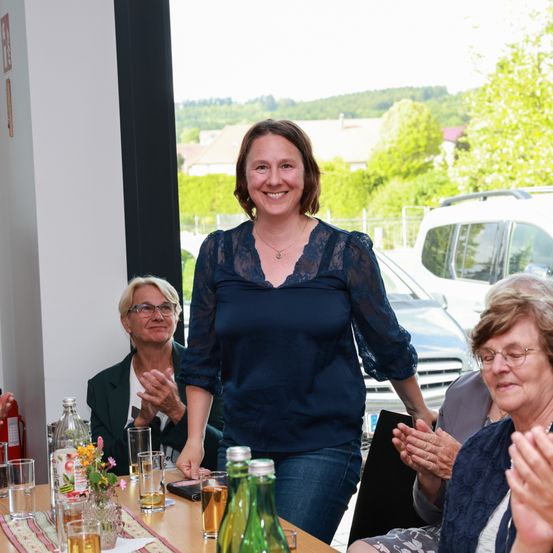 Eine Frau steht vor einem Tisch mit Getränken und lächelt, während andere klatschen. Dahinter klatschen zwei Frauen und ein Auto ist vor einer Glastür geparkt.