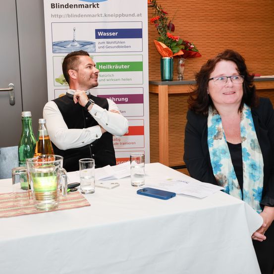 Ein Mann und eine Frau sitzen an einem Tisch mit verschiedenen Flaschen, einem Handy und Papieren. Dahinter bewirbt ein Schild 'Blindenmarkt' mit Optionen wie 'Wasser' und 'Heilkräuter.' Im Hintergrund befinden sich Blumen und ein Banner.