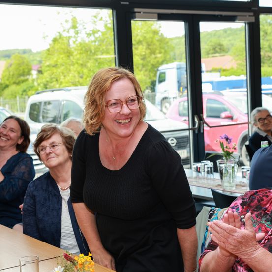 Eine Gruppe von Frauen sitzt um einen Tisch herum im Innenbereich und lächelt. Eine Frau mit Brille steht vor ihnen und lächelt. Draußen sind Autos geparkt, und Bäume sind sichtbar.