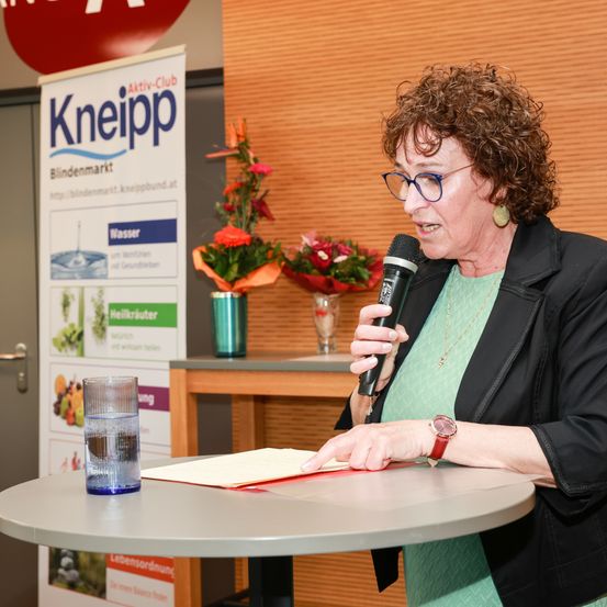 Eine ältere Frau mit Brille spricht in ein Mikrofon an einem Tisch, mit einem Kneipp-Banner und Blumen im Hintergrund.