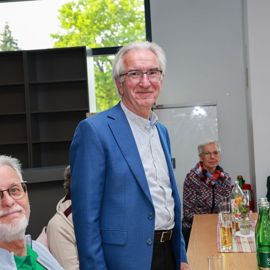 Ein Mann in einem blauen Sakko steht vor einem Tisch mit Getränken. Hinter ihm sitzen zwei ältere Personen, von denen eine eine Brille trägt. Der Raum ist innen, mit Bäumen, die durch ein Glasfenster zu sehen sind.