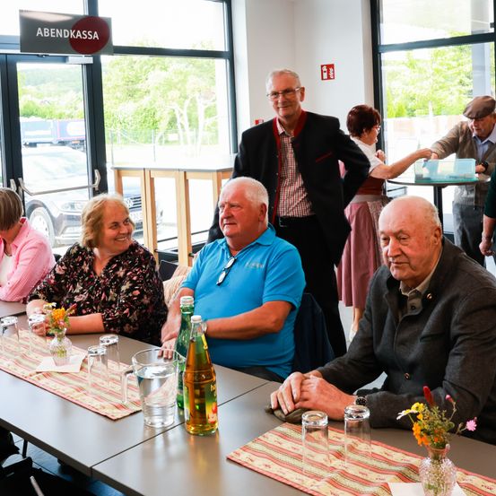 Eine Gruppe von Menschen sitzt an einem Tisch in einem Essbereich. Ein Mann in einem blauen Shirt und eine Frau mit Blumen in einer Vase sind unter ihnen. Das Schild oben steht 'ABENDKASSA'.