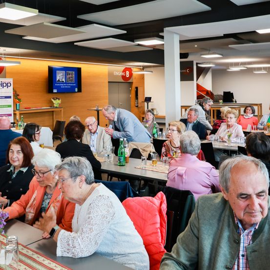 Eine Indoor-Versammlung von älteren Menschen an Tischen, mit einem Fernsehbildschirm im Hintergrund. Der Raum ist gut beleuchtet mit Deckenleuchten und einer hellen Atmosphäre.