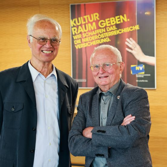 Zwei ältere Männer stehen nebeneinander und lächeln. Dahinter befindet sich ein Poster mit roten Vorhängen und deutschem Text.