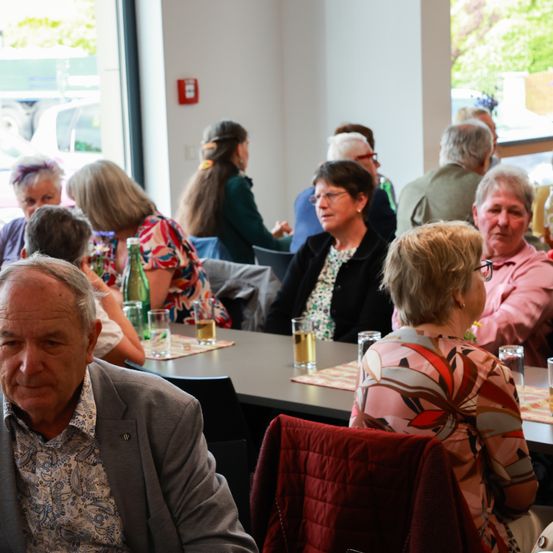 Eine Gruppe von Menschen sitzt um einen Tisch in einem Restaurant und führt Gespräche. Der Tisch ist mit Gläsern und Flaschen geschmückt. Draußen ist ein Auto am Fenster zu sehen.
