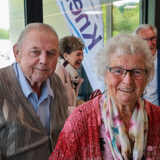 Ein älterer Mann und eine ältere Frau stehen nebeneinander und lächeln in die Kamera. Sie stehen vor einem Banner mit dem Wort 'Kern'. Dahinter steht eine Frau in einem Schal.