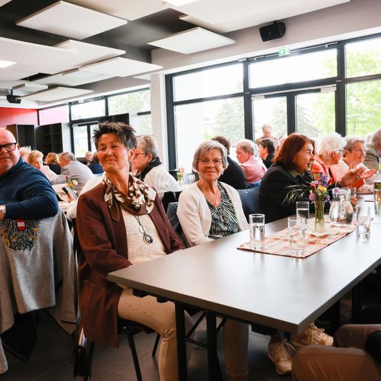 Mehrere Menschen sitzen um einen Tisch in einem Raum mit großen Fenstern. Zwei Frauen vorne tragen Brillen und Schals. Der Tisch hat Gläser, eine Vase und einen Tischset.