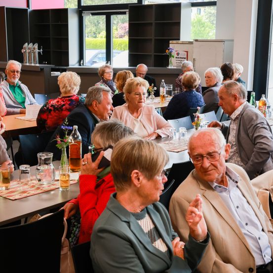 Eine Gruppe älterer Personen sitzt an einem Tisch in einem Speisebereich, einige unterhalten sich.