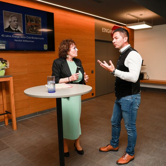 Ein Mann und eine Frau stehen in einem Raum. Der Mann spricht, während die Frau ein Mikrofon hält. Ein Tisch mit einem Glas steht zwischen ihnen. Ein Monitor ist an der Wand montiert.