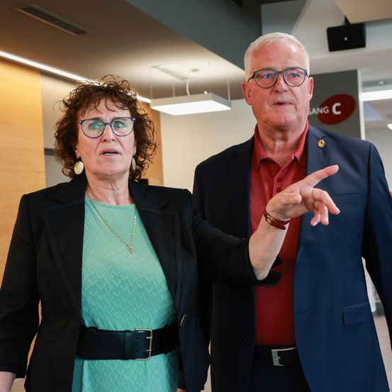 Eine ältere Frau in einem türkisfarbenen Kleid und ein Mann in einem roten Hemd und einem blauen Blazer zeigen auf etwas. Sie stehen in einem modernen Büroflur.