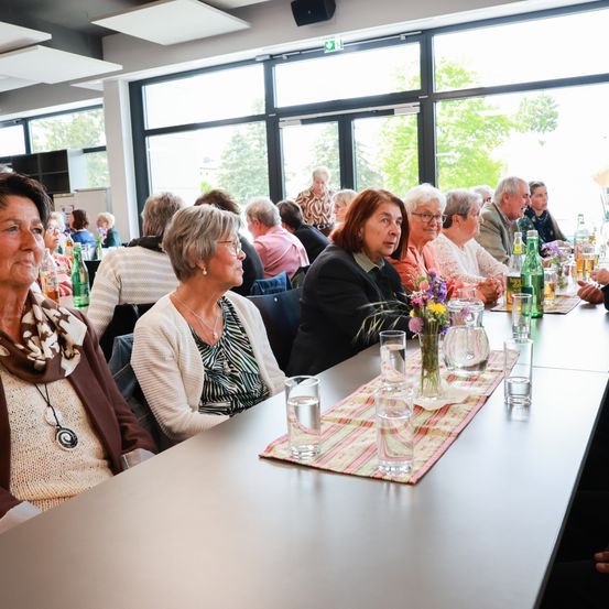 Eine Gruppe von Menschen sitzt an einem langen Tisch in einem Raum mit großen Fenstern. Sie essen, mit Gläsern und Blumen auf dem Tisch.