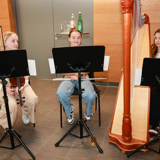 Drei junge Mädchen sitzen auf Stühlen und halten Musikinstrumente. Ein Mädchen hat eine Geige, ein anderes eine Flöte und das dritte eine Harfe. Sie scheinen sich in einem Musikzimmer zu befinden.