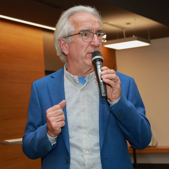 Ein älterer Mann in einem blauen Anzug mit Brille hält ein Mikrofon und gestikuliert, während er spricht. Dahinter ist eine Holzwand und eine Deckenleuchte.