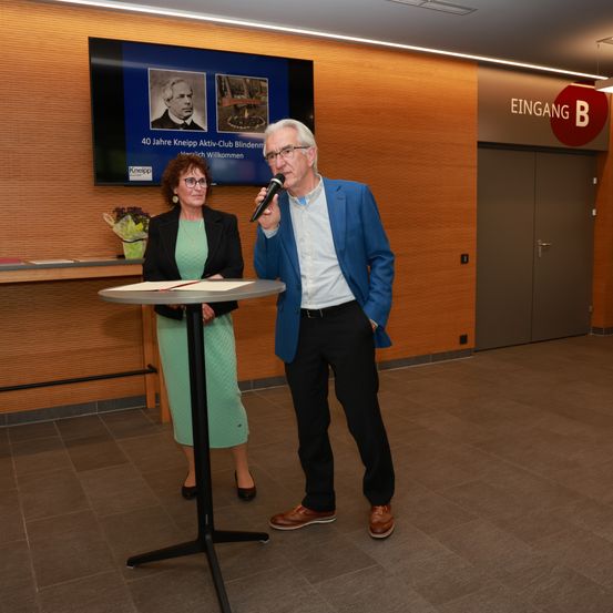 Zwei Personen, ein Mann und eine Frau, stehen an einem Tisch. Der Mann spricht in ein Mikrofon. Sie befinden sich in einem Raum mit Holzwänden und einem gefliesten Boden.