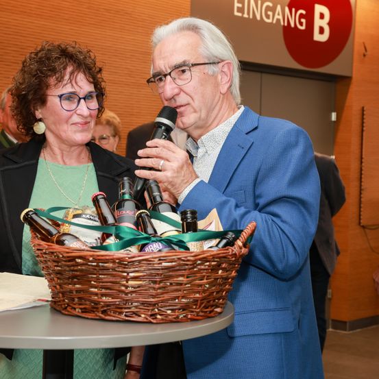 Ein älterer Mann hält ein Mikrofon und steht neben einer Frau, die einen Korb mit Flaschen hält und lächelt. Sie stehen vor einer Holzwand mit einem Schild, das 'EINGANG B' steht.