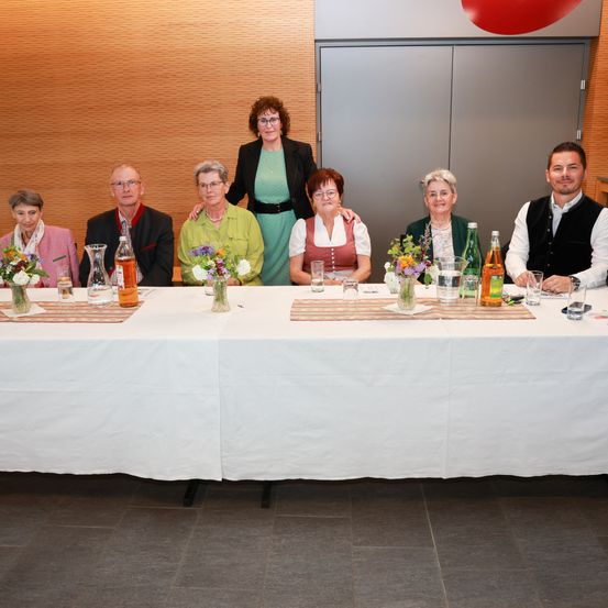 Eine Gruppe älterer Menschen, darunter Männer und Frauen, sitzen um einen Tisch mit Blumen, Gläsern und Flaschen. Sie posieren für ein Foto.