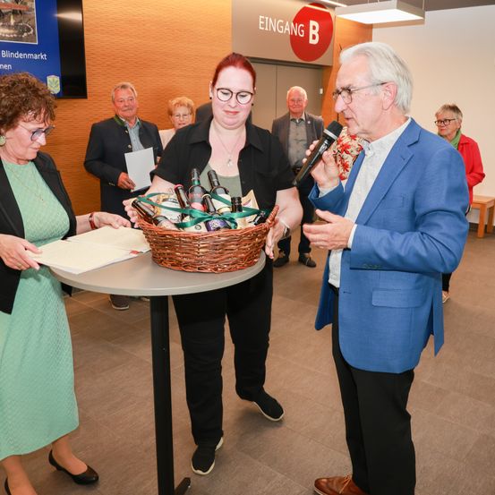 Eine Veranstaltung am Eingang eines Gebäudes. Ein Mann in einem blauen Anzug spricht in ein Mikrofon, während eine Frau in einem schwarzen Shirt einen Korb mit Flaschen hält. Eine andere Frau in einem grünen Kleid hält ein Papier. Dahinter steht eine Gruppe von Menschen.