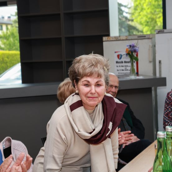 Eine ältere Frau mit einer Schürze lächelt und sitzt an einem Tisch. Hinter ihr sitzt ein Mann mit Brille am selben Tisch. Es gibt Regale und ein Schild mit Blumen auf der Theke.