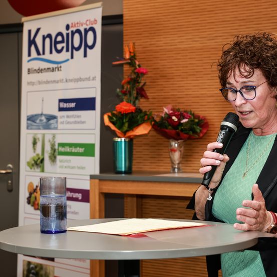 Eine Frau mit lockigem Haar und Brille spricht in ein Mikrofon, wahrscheinlich bei einer Veranstaltung. Ein Glas Wasser steht vor ihr auf einem Tisch. Ein Schild für Kneipp ist hinter ihr zu sehen.